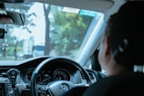 A person is seated in the driver's seat of a car, holding the steering wheel. The dashboard, including speedometers and other controls, is visible. Trees and a blurred view of vehicles can be seen through the windshield, indicating movement or driving. A small decoration is hanging from the rearview mirror.