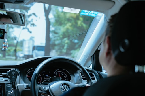 A person is seated in the driver's seat of a car, holding the steering wheel. The dashboard, including speedometers and other controls, is visible. Trees and a blurred view of vehicles can be seen through the windshield, indicating movement or driving. A small decoration is hanging from the rearview mirror.