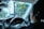 A person is seated in the driver's seat of a car, holding the steering wheel. The dashboard, including speedometers and other controls, is visible. Trees and a blurred view of vehicles can be seen through the windshield, indicating movement or driving. A small decoration is hanging from the rearview mirror.