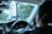 A person is seated in the driver's seat of a car, holding the steering wheel. The dashboard, including speedometers and other controls, is visible. Trees and a blurred view of vehicles can be seen through the windshield, indicating movement or driving. A small decoration is hanging from the rearview mirror.