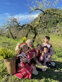 A group of children wearing colorful, traditional clothing are sitting on a grassy field surrounded by lush greenery and blooming trees. They appear to be enjoying a sunny day, with bright smiles and cheerful expressions. A basket filled with yellow flowers rests beside them, adding to the vibrant atmosphere.