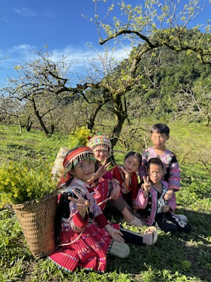 A group of children wearing colorful, traditional clothing are sitting on a grassy field surrounded by lush greenery and blooming trees. They appear to be enjoying a sunny day, with bright smiles and cheerful expressions. A basket filled with yellow flowers rests beside them, adding to the vibrant atmosphere.