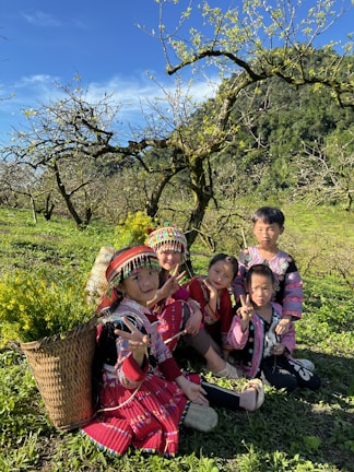 A group of children wearing colorful, traditional clothing are sitting on a grassy field surrounded by lush greenery and blooming trees. They appear to be enjoying a sunny day, with bright smiles and cheerful expressions. A basket filled with yellow flowers rests beside them, adding to the vibrant atmosphere.