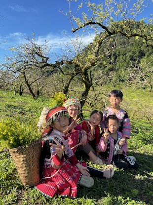 A group of children wearing colorful, traditional clothing are sitting on a grassy field surrounded by lush greenery and blooming trees. They appear to be enjoying a sunny day, with bright smiles and cheerful expressions. A basket filled with yellow flowers rests beside them, adding to the vibrant atmosphere.