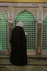 A person dressed in a black cloak with a white headscarf stands in front of an ornate silver grille. The grille is decorated with intricate patterns and features panels filled with green glass. Golden accents and Arabic script are visible above.
