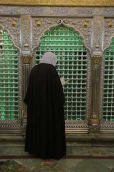 A person dressed in a black cloak with a white headscarf stands in front of an ornate silver grille. The grille is decorated with intricate patterns and features panels filled with green glass. Golden accents and Arabic script are visible above.