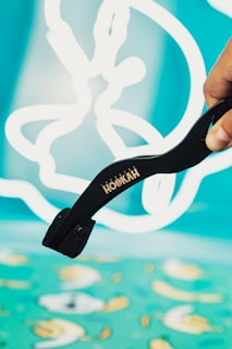 A hand is holding black tongs with the word 'HOOKAH' written on them. The background consists of abstract white shapes on a bright turquoise canvas, giving a playful and artistic ambiance.