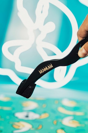 A hand is holding black tongs with the word 'HOOKAH' written on them. The background consists of abstract white shapes on a bright turquoise canvas, giving a playful and artistic ambiance.