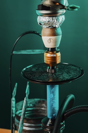 A close-up view of a hookah, featuring the bowl wrapped with aluminum foil and charcoal on top. The stem is blue and the base is connected to a hose. The background is a soft green, creating a relaxed atmosphere.