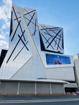 A modern architectural building with a geometric, glass facade featuring abstract black patterns. The structure includes an electronic billboard displaying an advertisement for Adidas Ultraboost Light shoes. The sky is clear and blue, enhancing the reflective quality of the building's surface.