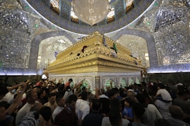 A crowd of people gathers around an ornate, gold-adorned shrine located inside a richly decorated room. The walls and ceiling are elaborately covered with mirrors, Arabic calligraphy, and blue and green embellishments. A large chandelier hangs in the center, enhancing the opulent atmosphere.