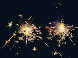 Two sparklers emitting bright, shooting sparks are held by a pair of hands. The background is dark, which enhances the glow and vibrancy of the sparks.