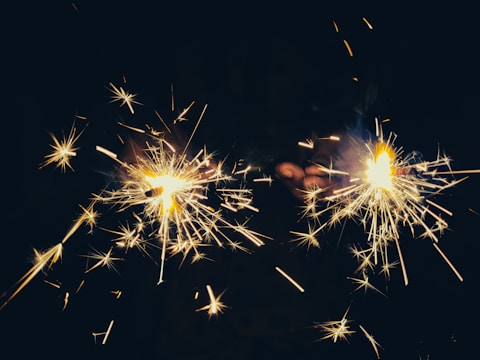 Two sparklers emitting bright, shooting sparks are held by a pair of hands. The background is dark, which enhances the glow and vibrancy of the sparks.