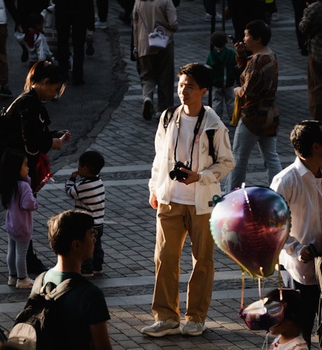 A young man holding a camera stands in the middle of a busy plaza, illuminated by a shaft of sunlight while the other people around him are in shadows. The scene includes adults and children, some holding balloons and engaging in various activities. The pavement is made of interlocking bricks and there's a sense of a vibrant, bustling public space.