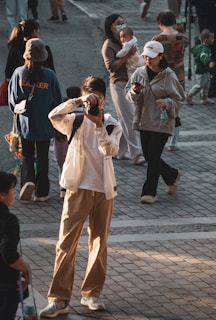 A busy street scene with a photographer capturing the moment, surrounded by people walking by. A person holds a baby, while another checks a smartphone. The lighting suggests early morning or late afternoon, with long shadows cast on the pavement.