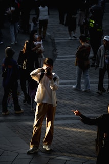 A person is standing in a crowd, taking a photograph with a camera. The scene is filled with people walking and engaging in various activities. The lighting highlights the individual with the camera, creating a strong contrast against the shaded background.