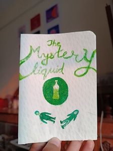 A hand is holding a white card with a textured pattern. The words 'The Mystery Liquid' are written in green cursive at the top. Below the text, there is a green circle containing the image of a bottle filled with a green liquid. Two green figure illustrations are shown lying down in a mirrored position below the circle.