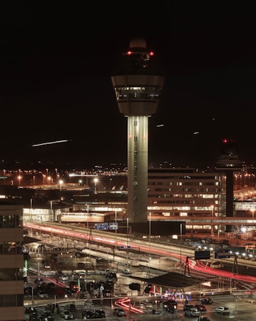 A control tower stands prominently at an airport during nighttime. Surrounding buildings are illuminated by artificial lights, and light streaks from vehicles are visible on the roads in the foreground, indicating movement. The sky is dark, enhancing the brightness of the lights.