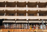 A beige multi-story building with rows of balconies and a ground-floor supermarket with striped awnings. The supermarket facade displays various items and colorful signage.