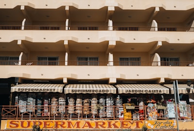 A beige multi-story building with rows of balconies and a ground-floor supermarket with striped awnings. The supermarket facade displays various items and colorful signage.