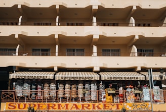 A beige multi-story building with rows of balconies and a ground-floor supermarket with striped awnings. The supermarket facade displays various items and colorful signage.