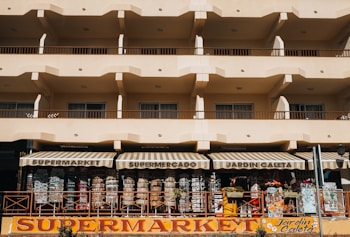 A beige multi-story building with rows of balconies and a ground-floor supermarket with striped awnings. The supermarket facade displays various items and colorful signage.
