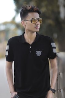 A person wearing sunglasses and a black polo shirt with a geometric animal design on the chest, standing outdoors near a railing with blurred greenery in the background.