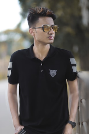 A person wearing sunglasses and a black polo shirt with a geometric animal design on the chest, standing outdoors near a railing with blurred greenery in the background.