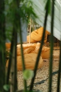 A cozy outdoor setting featuring several orange bean bags arranged on a bed of pebbles. The scene is framed by green plant stems or bamboo, adding a natural barrier. Sunlight filters through, casting a warm, inviting glow across the bean bags.