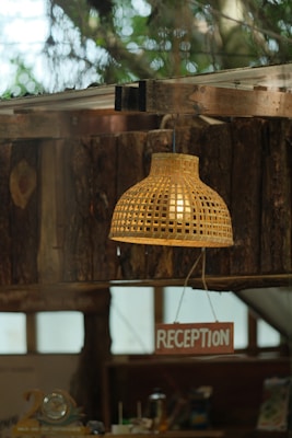 A woven basket-style lamp hangs from the ceiling, casting a warm glow above a wooden structure. Below the lamp, a sign labeled 'RECEPTION' is prominently displayed. The background features rustic wooden walls and blurred greenery outside, giving a cozy, natural ambiance.