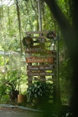 A wooden signpost surrounded by lush greenery, displaying several signs pointing towards a reception, restaurant, and glamping areas with lake and garden views. The signpost is decorated with circular wreaths and flanked by potted plants, set amidst a serene outdoor environment.
