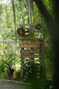 A wooden signpost surrounded by lush greenery, displaying several signs pointing towards a reception, restaurant, and glamping areas with lake and garden views. The signpost is decorated with circular wreaths and flanked by potted plants, set amidst a serene outdoor environment.