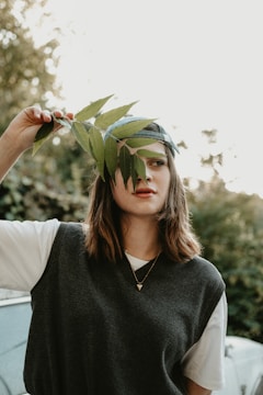 A woman with shoulder-length brown hair is holding a green branch in front of her face. She wears a dark sleeveless sweater over a white shirt and a necklace with a small heart pendant. The background is a blurred green, suggesting a natural setting.