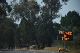 A mobile electronic traffic sign displays the message 'Prepare to Stop' on a road, surrounded by tall trees and vegetation. The scene appears to be captured during daylight with a slightly overcast sky.