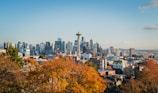 A vibrant cityscape featuring a skyline with numerous modern skyscrapers and the iconic Space Needle. In the foreground, there are trees with autumn-colored leaves, providing a contrast to the urban setting.