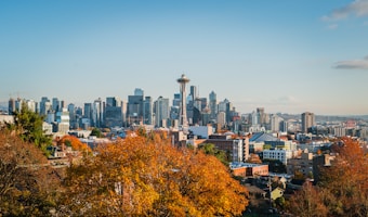 A vibrant cityscape featuring a skyline with numerous modern skyscrapers and the iconic Space Needle. In the foreground, there are trees with autumn-colored leaves, providing a contrast to the urban setting.