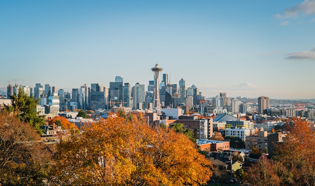 A vibrant cityscape featuring a skyline with numerous modern skyscrapers and the iconic Space Needle. In the foreground, there are trees with autumn-colored leaves, providing a contrast to the urban setting.