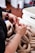 Hands are working with raw sausage links in a white container. The person has a tattoo on the arm and is wearing a maroon watch. There is some kitchen equipment and items in the background, including a large white container and other items out of focus.