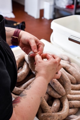 Hands are working with raw sausage links in a white container. The person has a tattoo on the arm and is wearing a maroon watch. There is some kitchen equipment and items in the background, including a large white container and other items out of focus.