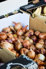 A pile of onions is stacked inside a cardboard box. The onions are brown with hints of purple and have dry, papery skins. In the background, there are more cardboard boxes, one labeled with 'Parsnips' containing white root vegetables.