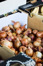 A pile of onions is stacked inside a cardboard box. The onions are brown with hints of purple and have dry, papery skins. In the background, there are more cardboard boxes, one labeled with 'Parsnips' containing white root vegetables.