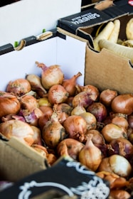 A pile of onions is stacked inside a cardboard box. The onions are brown with hints of purple and have dry, papery skins. In the background, there are more cardboard boxes, one labeled with 'Parsnips' containing white root vegetables.