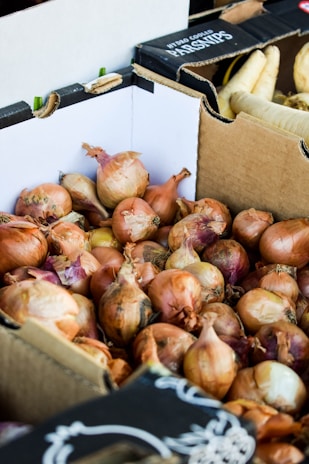 A pile of onions is stacked inside a cardboard box. The onions are brown with hints of purple and have dry, papery skins. In the background, there are more cardboard boxes, one labeled with 'Parsnips' containing white root vegetables.