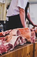 Chunks of raw meat are arranged on a wooden butcher's block. In the background, a person stands wearing a white shirt and dark apron, handling the meat.