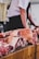 Chunks of raw meat are arranged on a wooden butcher's block. In the background, a person stands wearing a white shirt and dark apron, handling the meat.