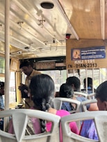Interior of a bus with several passengers seated, a conductor standing and checking tickets. The roof of the bus is equipped with handles for standing passengers. A signboard with text and an image is visible, along with some small framed pictures of deities or notable figures.