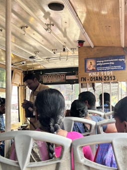 Interior of a bus with several passengers seated, a conductor standing and checking tickets. The roof of the bus is equipped with handles for standing passengers. A signboard with text and an image is visible, along with some small framed pictures of deities or notable figures.