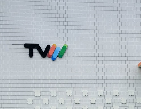 A white tiled wall features a logo consisting of the letters 'TV' followed by four curved lines in red, blue, green, and yellow. Below the logo, there are decorative elements shaped like bear faces aligned in rows. The bottom right corner shows a partial view of an orange object.