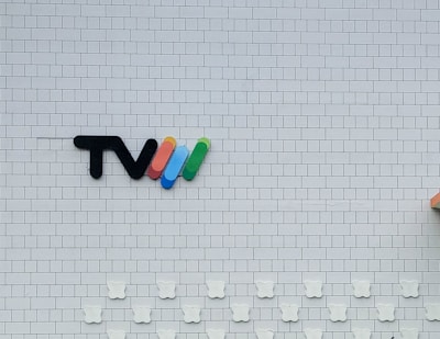 A white tiled wall features a logo consisting of the letters 'TV' followed by four curved lines in red, blue, green, and yellow. Below the logo, there are decorative elements shaped like bear faces aligned in rows. The bottom right corner shows a partial view of an orange object.