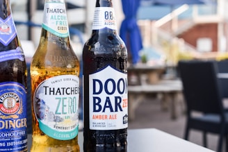 Three bottles of non-alcoholic beverages are arranged on a table in an outdoor setting. The bottles are branded as Erdinger Weissbrau, Thatchers Zero, and Doom Bar Zero. The background features blurred patio furniture and blue umbrellas, suggesting a relaxed and casual environment.